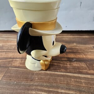 May include: A vintage Mickey Mouse figurine wearing a cream-colored top hat with a gold band. The figurine has black ears, a black nose, and a gold bow tie. The figure is made of plastic and is on a wooden surface.