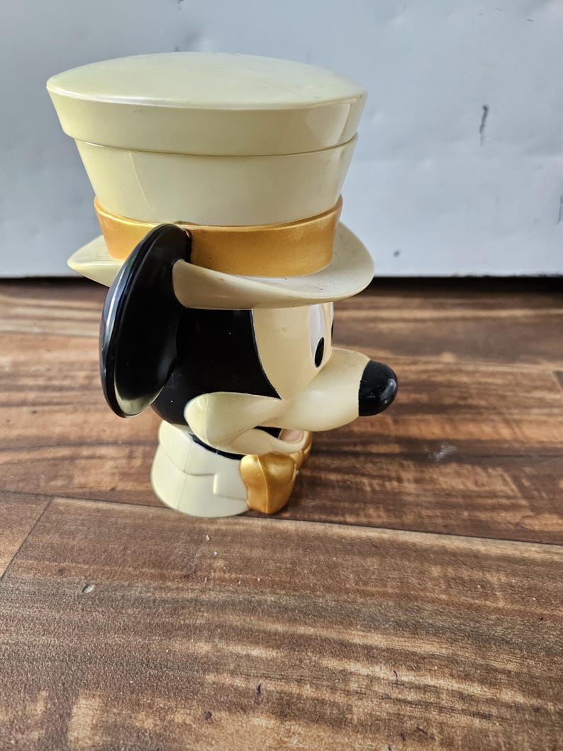May include: A vintage Mickey Mouse figurine wearing a cream-colored top hat with a gold band. The figurine has black ears, a black nose, and a gold bow tie. The figure is made of plastic and is on a wooden surface.