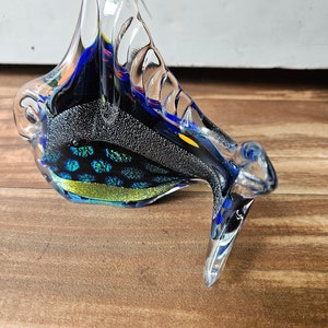 May include: A glass sculpture of a fish with blue, yellow, and black accents. The fish has a clear glass body with a textured surface.