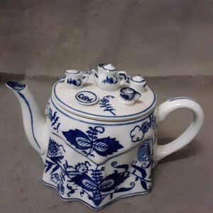 May include: A white teapot with a blue and white floral design. The teapot has a lid that is shaped like a table with miniature teacups and a teapot on top.