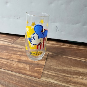 May include: A clear glass drinking glass with a Mickey Mouse design. The glass has a yellow background with red and blue stripes and yellow stars. The text "mickey" is printed on the bottom of the glass.