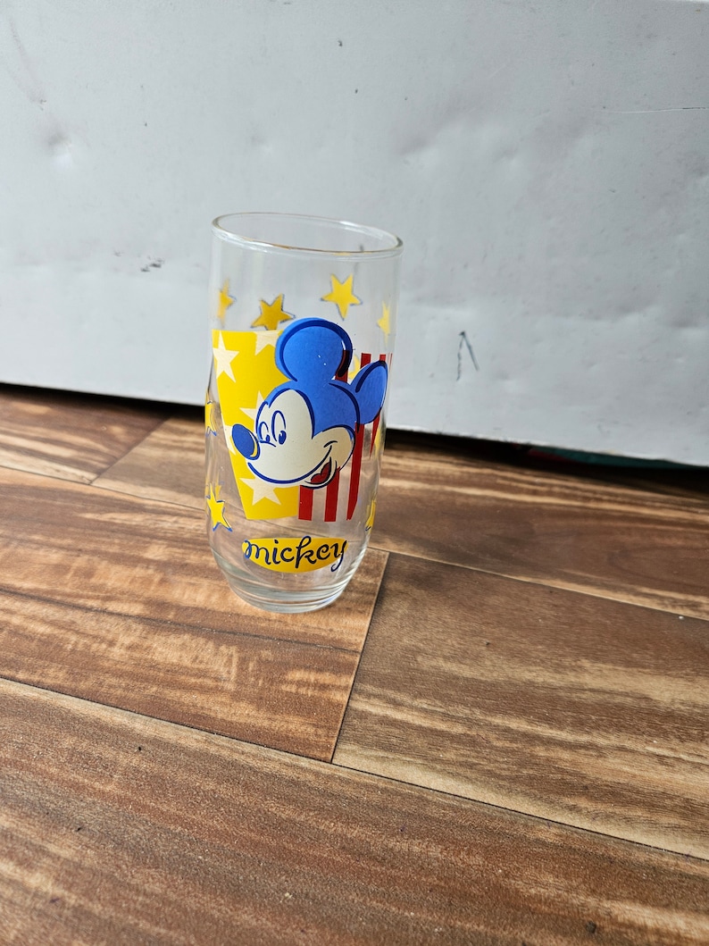 May include: A clear glass drinking glass with a Mickey Mouse design. The glass has a yellow background with red and blue stripes and yellow stars. The text "mickey" is printed on the bottom of the glass.