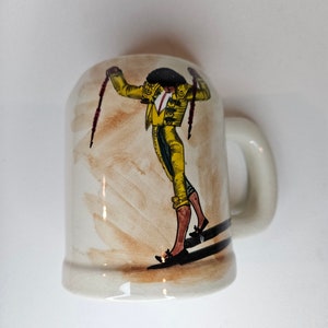 May include: White ceramic mug with a brown and yellow illustration of a matador holding a cape. The matador is wearing a yellow and green suit with a red sash.