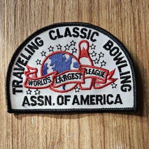 May include: White embroidered patch with black trim. It says "Traveling Classic Bowling Assn. of America" with a red, white, and blue globe and bowling pin graphic. The globe has the words "World's Largest League" on a red ribbon.