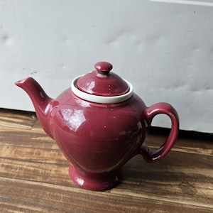 May include: A burgundy ceramic teapot with a white lid and a white rim around the lid. The teapot has a rounded body and a spout on one side and a handle on the other.