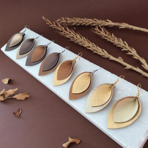 Puede incluir: Una colección de pendientes en forma de hoja en capas en varios tonos de marrón y dorado. Los pendientes se muestran sobre una superficie rectangular blanca sobre un fondo marrón. Cada pendiente tiene un gancho plateado u dorado.