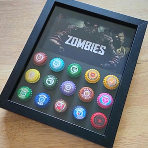 May include: A black rectangular shadow box frame containing a graphic print with the word ZOMBIES and fourteen colorful bottle caps arranged in a grid. Each bottle cap features a unique circular icon and text, including colors like red, blue, green, yellow, purple, and brown.