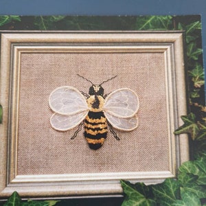 May include: A framed embroidery of a bee. The bee features a black and yellow striped body, delicate white wings, and black antennae. The embroidery is set against a tan fabric background within a light-coloured wooden frame. Green ivy surrounds the frame.