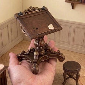 May include: A miniature wooden writing desk with a slanted top, featuring ornate gold-toned carved details on the base and edges. The desk includes two small inkwells, a pen, and a tiny piece of paper. A matching small round stool is positioned nearby.