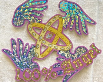 May include: A pink and gold sticker with a design of a circle with eyes and wings. The text "100% Angel" is written on the sticker.