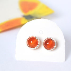 以下が含まれることがあります： オレンジのカーネリアン宝石のイヤリングのペア。シルバーワイヤーのセッティングが施されています。イヤリングは白いカードに飾られています。