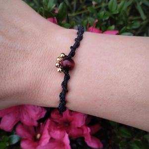 May include: Black macramé bracelet with a central red-brown bead and small gold-coloured beads. The bracelet is displayed on a wrist, with pink flowers and green foliage in the background.
