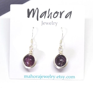 Puede incluir: Un par de pendientes de plata con cuentas de piedras preciosas moradas. Los pendientes están envueltos en alambre y tienen un cierre de gancho simple. La tarjeta dice "Mahora Jewelry" y "mahorajewelry.etsy.com".