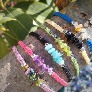 Könnte beinhalten: Eine Sammlung bunter Armbänder mit verschiedenen farbigen Schnüren und Edelsteinperlen. Die Armbänder sind auf einem Baumzweig angeordnet.