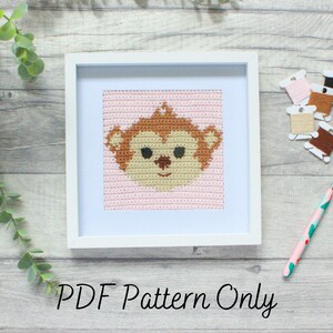May include: A white frame with a pink and brown crocheted monkey face inside. The frame is on a woodgrain surface with a crochet hook and yarn spools.