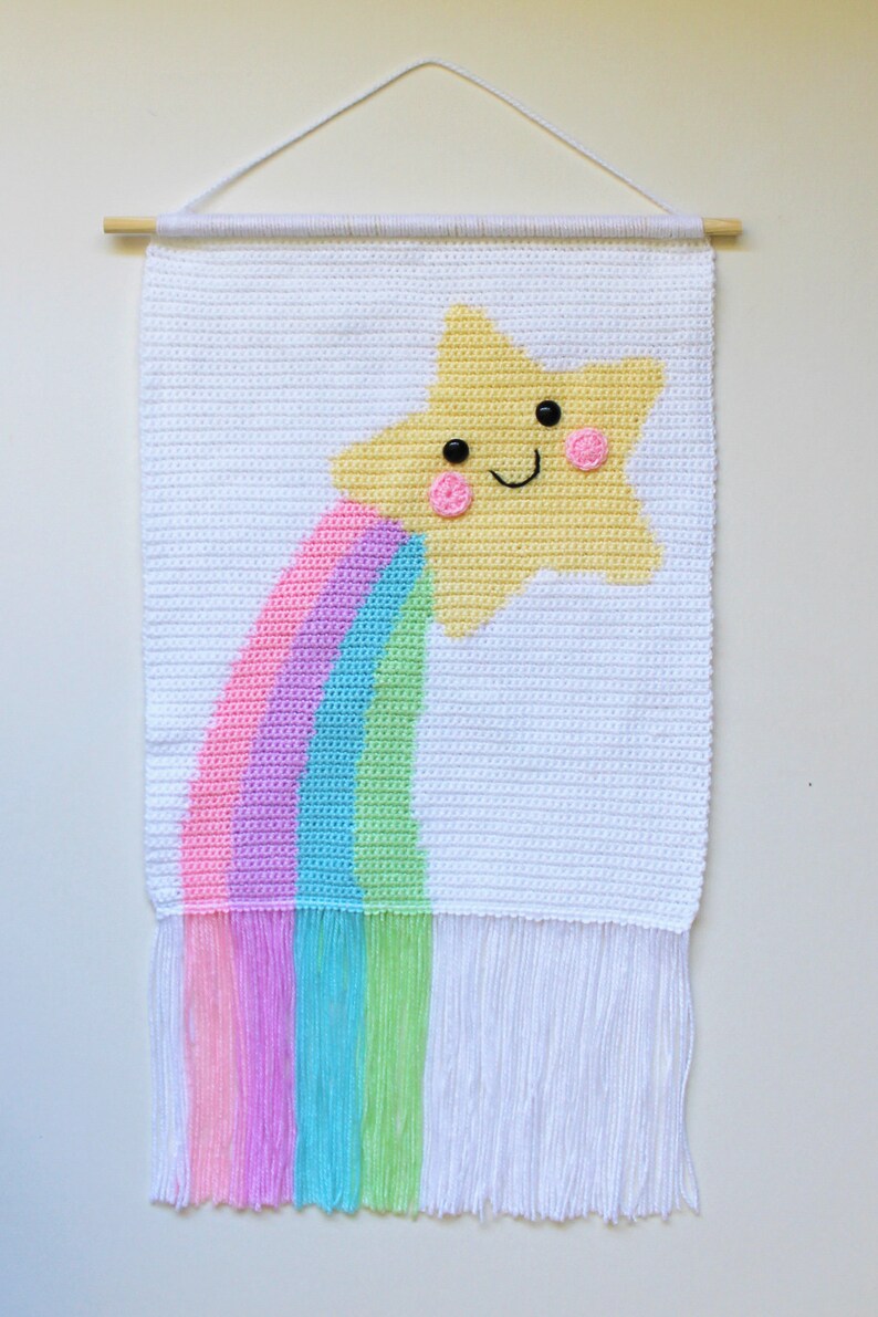 May include: A white crocheted wall hanging with a yellow star and a rainbow. The star has a smiling face with black eyes and pink cheeks. The rainbow is made of pink, purple, light blue, and green yarn. The wall hanging has white fringe.