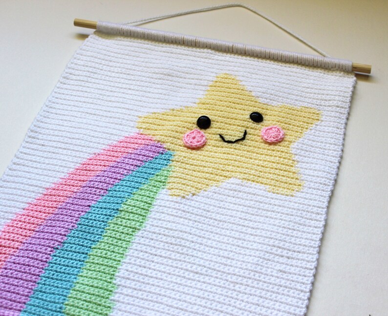 May include: A white crocheted wall hanging with a yellow star and a rainbow. The star has a smiling face with black eyes and pink cheeks.