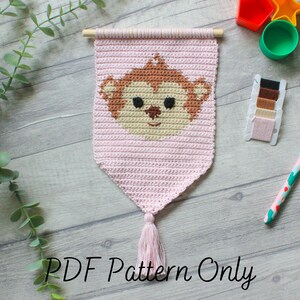 Könnte beinhalten: Ein rosa gehäkelter Wandbehang mit einem braunen Affengesicht in der Mitte. Der Behang hat unten einen Quasten und ist aus verschiedenenfarbigen Garnen gefertigt. Der Text "PDF Pattern Only" steht unter dem Behang.