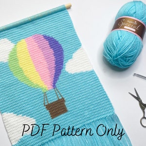 May include: A crochet pattern for a hot air balloon wall hanging. The pattern features a blue sky background with white clouds and a rainbow coloured hot air balloon. The pattern is available as a PDF download only. The image also shows a ball of light blue yarn, a crochet hook, and scissors.