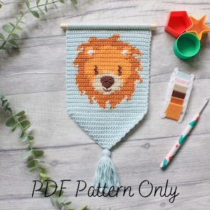 May include: Crochet pattern for a lion head wall hanging. The pattern includes a light blue background with a brown lion head and a light blue tassel. The pattern is available as a PDF download.