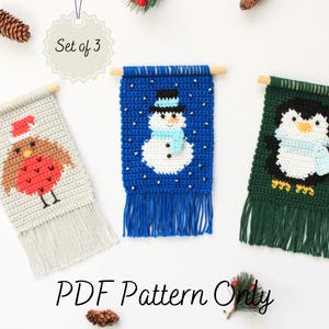 May include: A set of three crochet wall hangings featuring a snowman, a penguin, and a robin. Each hanging is made with colorful yarn and has a fringe at the bottom. The text "Set of 3" is visible in the image.