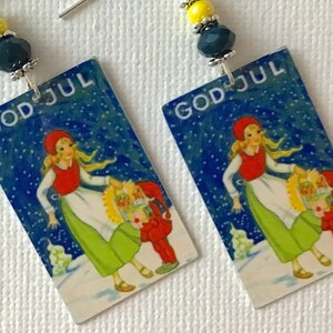 May include: A pair of earrings featuring a vintage Christmas card image with the text "God Jul" in blue and white. The card depicts a girl in a red hat and green skirt with a small child in a red suit. The earrings are accented with blue and yellow beads.
