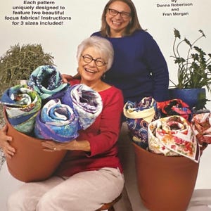 May include: A book titled "Double Focus 3-Yard Quilts" by Donna Robertson and Fran Morgan. The cover features two women holding rolled up quilts in terracotta pots. The book cover is white with blue and purple text.