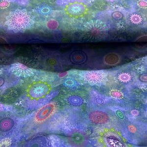 May include: Close-up of fabric with a vibrant, colorful floral pattern. The design features mandala-like shapes in shades of purple, pink, green, and orange against a blue background. The fabric has a textured appearance.