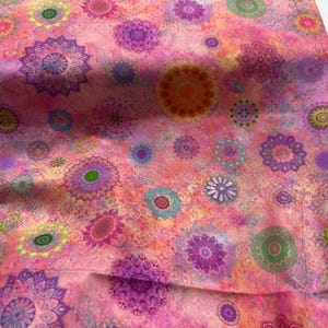 May include: Pink fabric with a repeating pattern of colorful mandala designs in shades of purple, green, orange, and blue. The fabric has a watercolor effect, with a soft, textured appearance. Ideal for quilting or crafting projects.