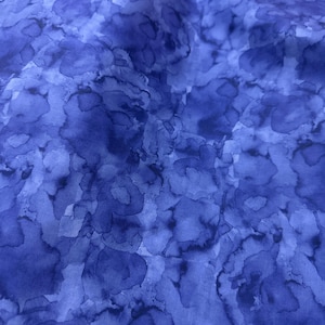 May include: Close-up of a fabric with a watercolor-like pattern. The fabric features a blend of blue hues, with darker shades creating a mottled effect against a lighter blue background. The design resembles abstract shapes.