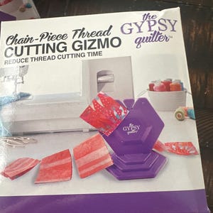 May include: A purple Chain-Piece Thread Cutting Gizmo by The Gypsy Quilter. The box features the product name and the phrase "Reduce Thread Cutting Time." The image shows the gizmo with cut fabric pieces in red and white.