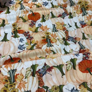 May include: A fabric pattern featuring a dense arrangement of white and orange pumpkins interspersed with colorful autumn leaves, small yellow flowers, and dark green stems. The design creates a repetitive seasonal motif with a light background.