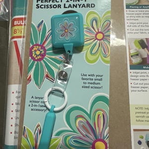 May include: A teal Perfect 2-in-1 scissor lanyard with a floral design. The lanyard is designed to hold small to medium-sized scissors. The packaging features a floral pattern in teal, green, pink, and yellow.