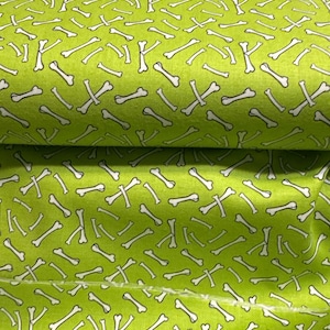 May include: Bright green fabric with a repeating pattern of white and black dog bones. The bones are scattered across the fabric, creating a playful and whimsical design. The fabric is folded, showing the texture and depth of the material.