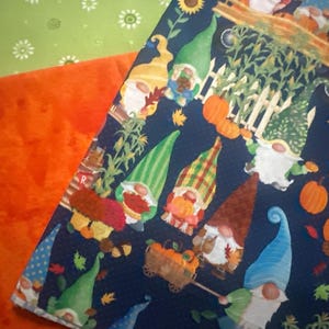 May include: A dark blue fabric featuring a repeating pattern of gnomes in various hats, pumpkins, corn stalks, and sunflowers. The gnomes are depicted with white beards and colorful clothing. The fabric is layered over solid orange and light green fabric.