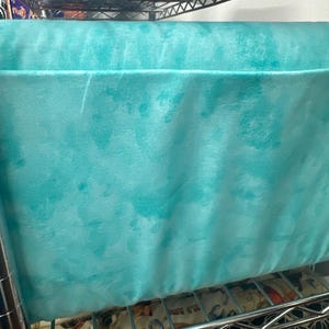 May include: A large, rectangular, turquoise fabric item with a tie-dye effect, resting on a metal wire shelf. The fabric has a soft, textured appearance. The shelf below contains patterned fabric.