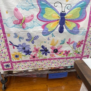 May include: A white fabric panel featuring a colorful butterfly and floral design. The butterflies have blue, pink, yellow, and purple wings. The border has a pink heart pattern. The fabric is on a metal rolling cart.