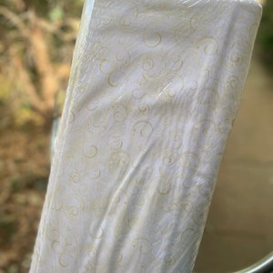 May include: A roll of white fabric with a subtle swirl pattern in a light gold color. The fabric is wrapped in clear plastic.