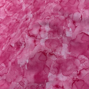 May include: Close-up of a pink fabric with a watercolor-like pattern. The design features overlapping, irregular shapes in varying shades of pink and magenta, creating a textured, artistic effect. The fabric appears soft and flowing.