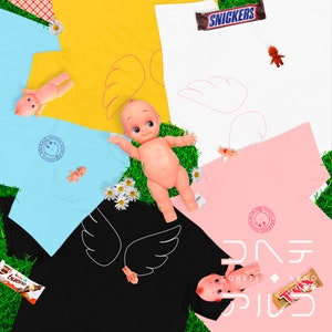 May include: Five different t-shirts with a variety of colors and designs. Each shirt features a pair of wings and a small doll. The shirts are laid out on a green grassy surface with candy bars and flowers.