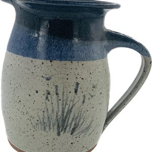 May include: A ceramic pitcher with a dark blue rim and handle, transitioning to a lighter blue and speckled gray body. The pitcher features a hand-painted design of reeds and grasses. The base is a warm brown.