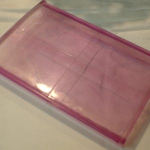 May include: A rectangular, transparent pink plastic container with a raised pink border. The container has a slightly curved shape and appears to be empty. The surface has some minor imperfections.