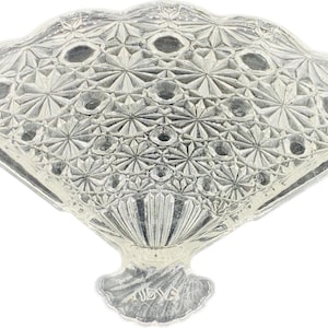 May include: A silver-colored, fan-shaped decorative dish with a detailed, geometric pattern. The dish has a scalloped edge and a textured surface, with a base that resembles a stylized flower. The fan design is symmetrical and ornate.
