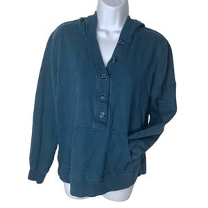 May include: A teal-colored hooded sweatshirt with a V-neckline and a button-up closure. The sweatshirt has a kangaroo pocket and long sleeves. The garment is displayed on a white mannequin.