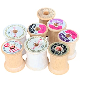 May include: A collection of vintage wooden thread spools. The spools are various sizes and feature colorful labels with text and designs. The spools are made of light-colored wood and are arranged on a white background.