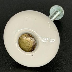May include: A round, off-white ceramic cabinet knob featuring a smooth, glossy finish and a centred, circular brass-toned metal inlay. The knob includes a threaded silver-coloured metal bolt extending from the back for secure installation.