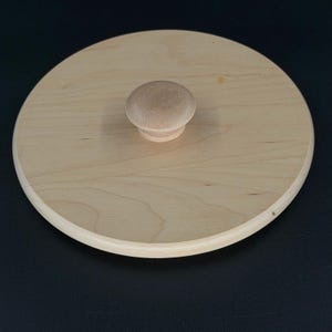 Puede incluir: Una tapa circular de madera de color claro con una superficie lisa y plana que muestra un patrón de veta natural. Un pequeño pomo de madera redondeado está centrado en la parte superior, sirviendo como asa para levantar la tapa.