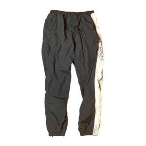 May include: Black and beige athletic pants with the brand name "Reebok" printed vertically on the right leg. The pants have an elastic waistband and cuffs at the ankles. The material appears to be a lightweight nylon or polyester.