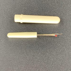 May include: Ivory seam ripper with a protective cap. The tool has a metal blade with a red ball at the tip. The cap is cylindrical and fits over the handle. The seam ripper is designed for removing stitches from fabric.