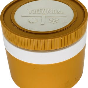 May include: A vintage Thermos container with a mustard yellow base and lid, separated by a white band. The lid features the word "THERMOS" in white, along with snowflake and sunburst designs.